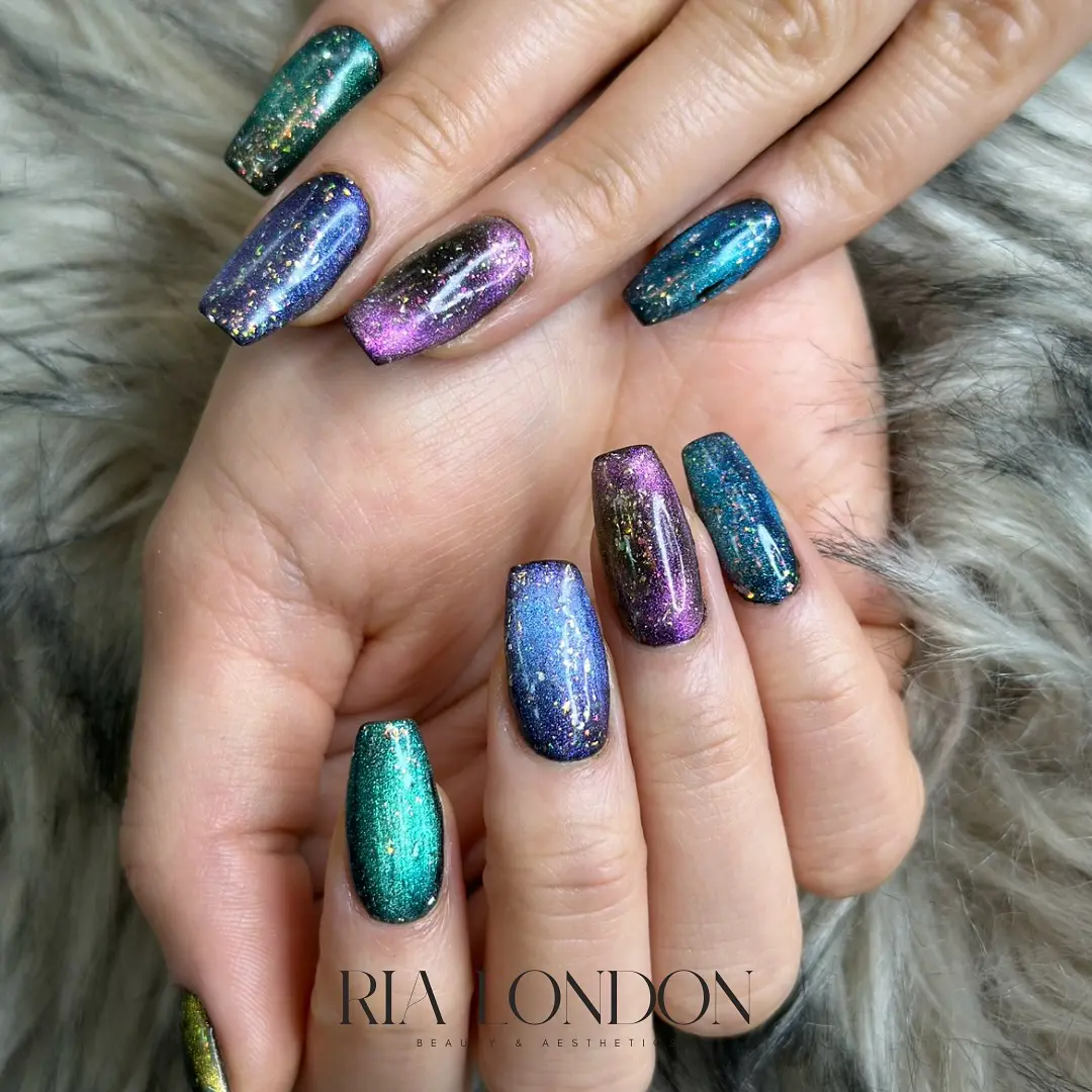 A set of manicured nails with a multicolored, galaxy-inspired design featuring shimmering blues, purples, greens, and gold accents. The nails are displayed on a soft, gray, textured background.