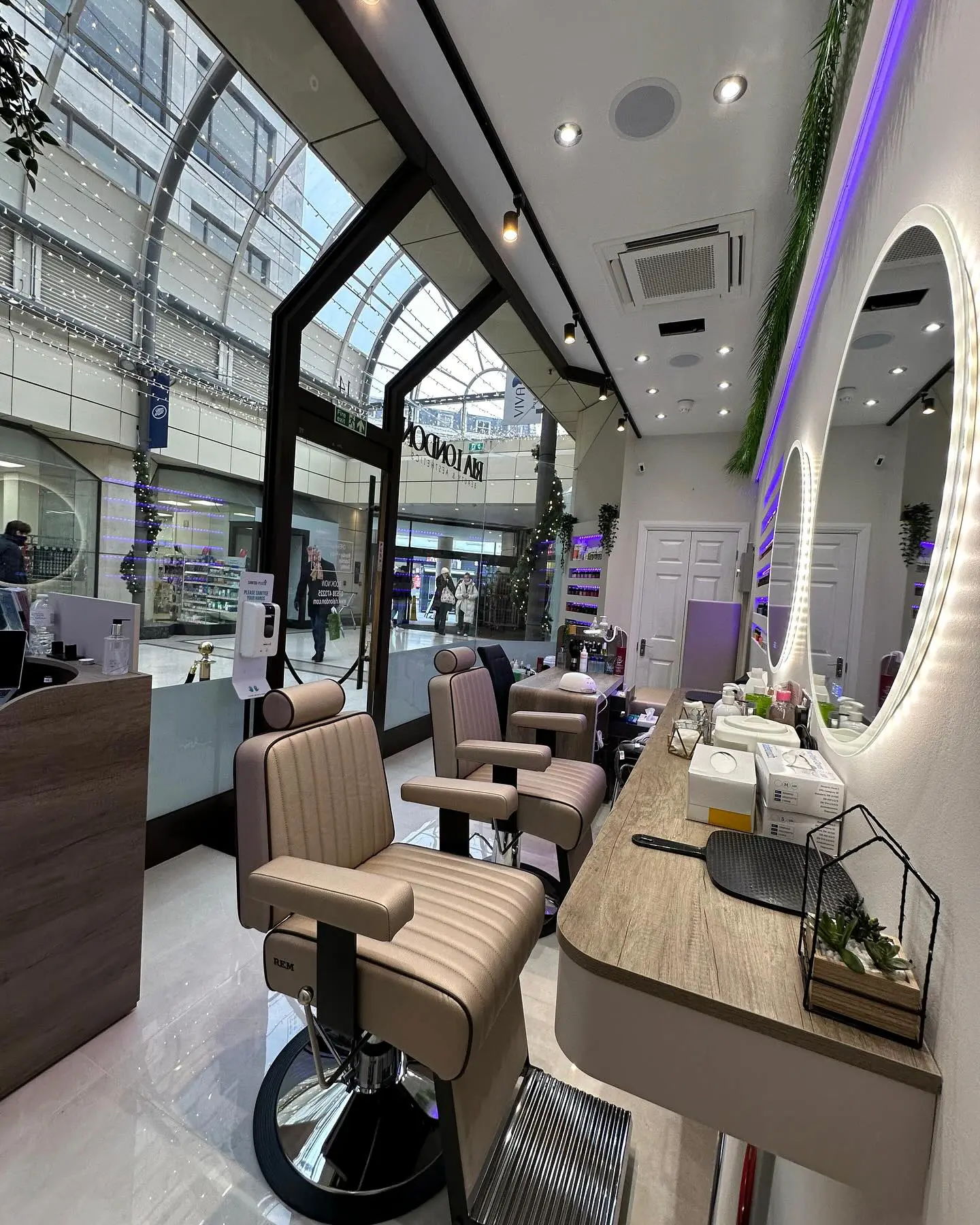 A modern barbershop interior with sleek beige barber chairs, large circular mirrors with LED lighting, and a glass facade showing a view of a shopping mall. The salon has a clean, minimalistic design with white walls, greenery decor, and accent lighting.