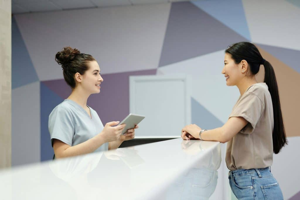 A smiling receptionist in medical scrubs assisting a woman at a clinic reception desk with a tablet in hand.